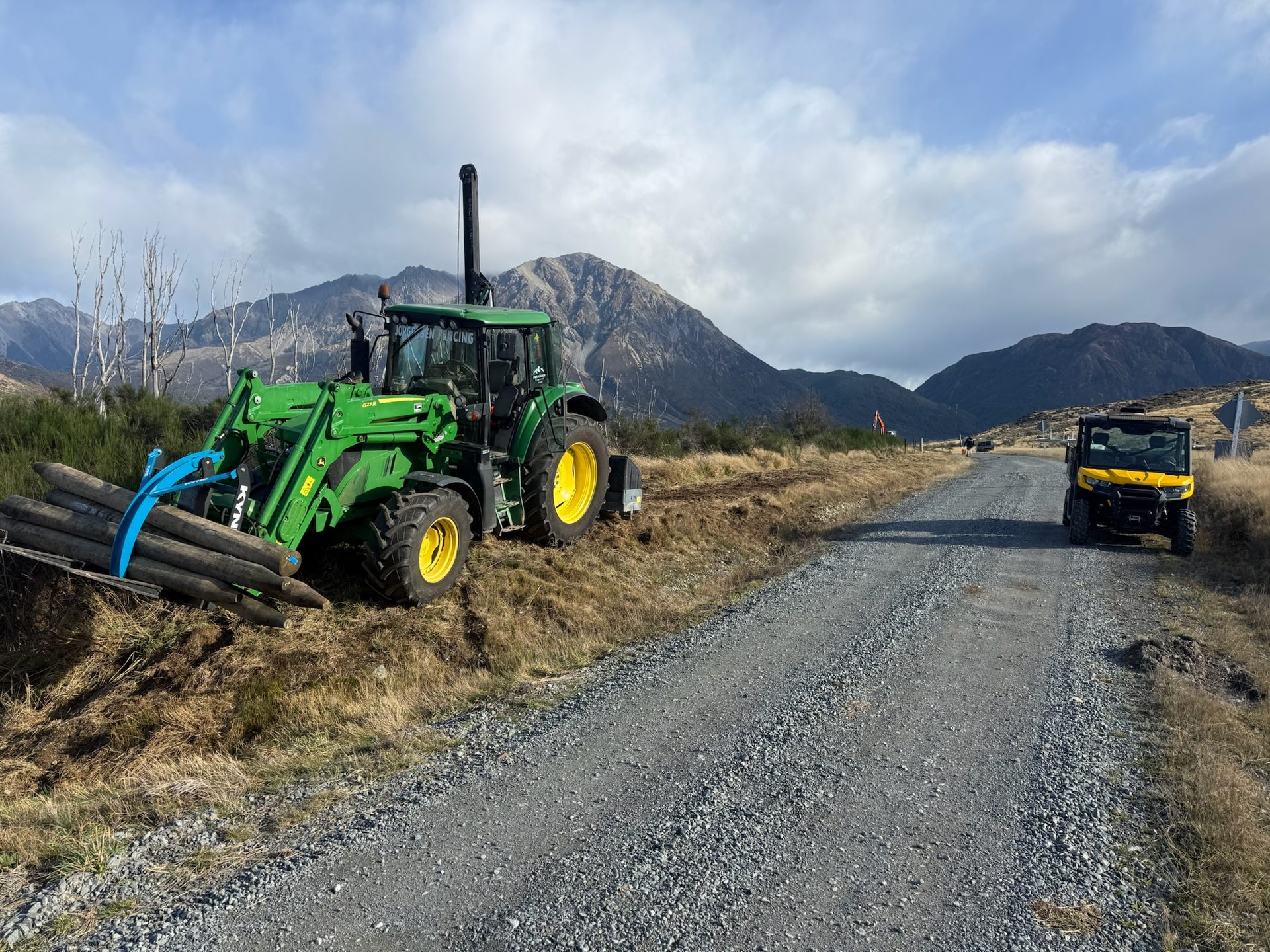 Jorgensen Fencing tractor loaded with timber posts on a gravel high-country road, Canterbury Southern Alps behind